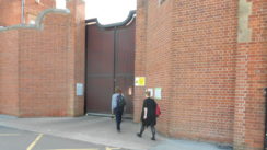 Picture of two people walking in a prison entrance, a large gate next to a red brick wall