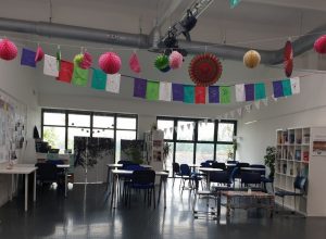 Photograph of a large room with tables and chairs arranged in various formations, a bookshelf, large windows to the back and bunting hanging from the ceiling.